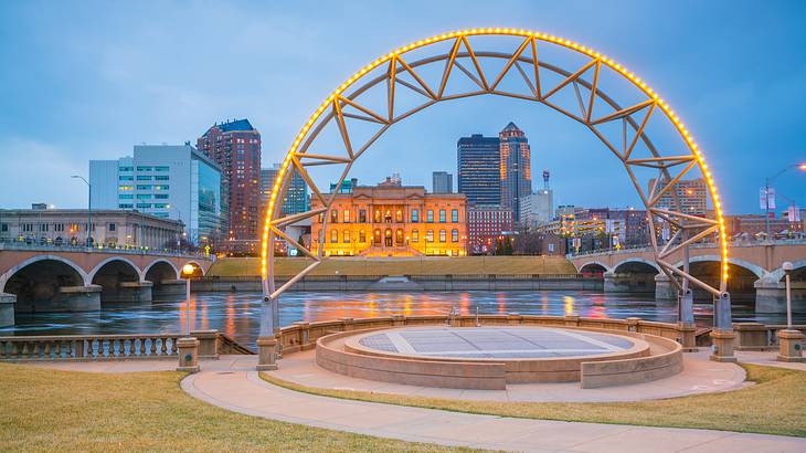 A city skyline from a river bank with a lit-up arch during twilight hours